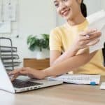 High school woman doing paperwork at desk with laptop.