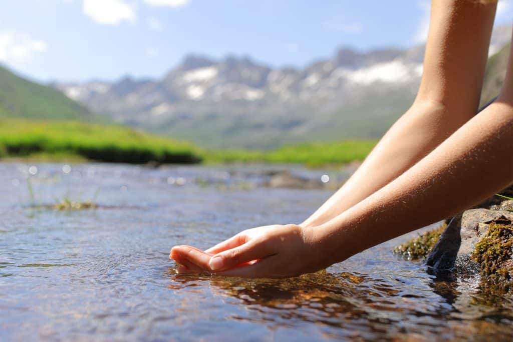 Hands touching flowing river water in scenic mountain landscape.